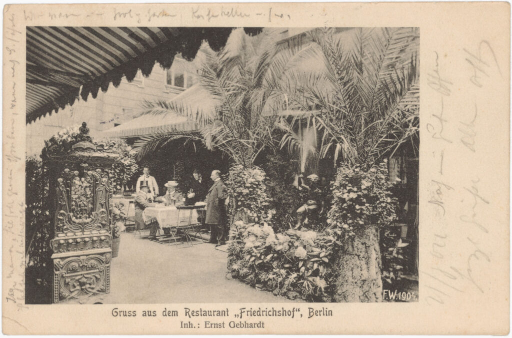 Vorne links im Bild ist ein Süßwarenautomat der Firma „Hartwig & Vogel“ zu sehen. In den hier käuflichen Schokoladen waren vermutlich auch kolonialrassistische Sammelbilder enthalten. Innenhof vom Restaurant “Friedrichshof”, Friedrichstr., o.D. (verschickt am 21.08.1904), SPP / FHXB 0965